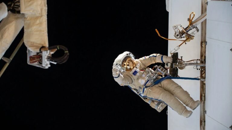 From Soyuz to the stars: A Roscosmos trivia quiz an astronaut in a white space suit hangs on to a metal bar on the outside of a large white structure. the blackness of space fills the background