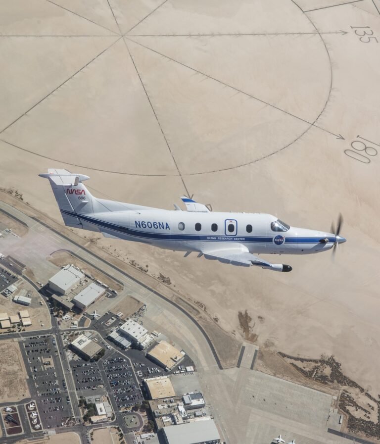 NASA’s freshly painted Pilatus PC-12 flies over NASA’s Armstrong Flight Research Center in Edwards, California. The parking lots and center buildings dot the landscape below. The compass rose in the upper part of the photo shaped like the sun hovers over the aircraft, emphasizing the gleaming white plane with a blue stripe and blue N606NA number across the fuselage and NASA red worm logo on the tail. A sensor, with a black-tip juts out from below the wing.