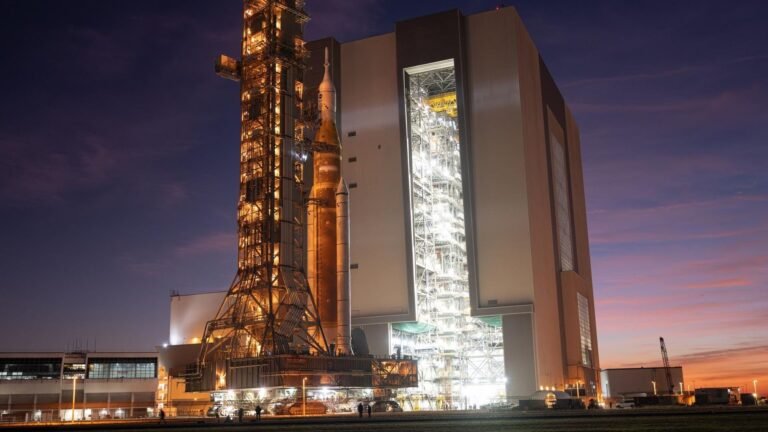 NASA repairs Artemis 2 rocket, continues eyeing April moon launch a large orange rocket beside metal scaffolding on a wheeled platform rolls into a massive hangar building in front of a purple and orange sunset