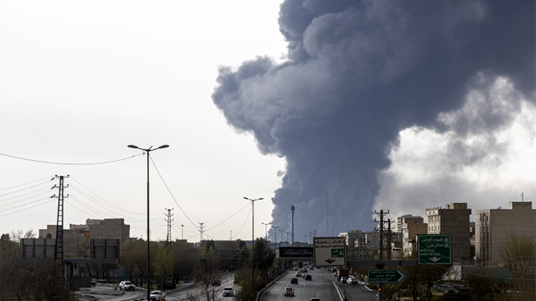 Cars drive down a highway as smoke billows after overnight air strikes on oil depots on March 8, 2026 in Tehran, Iran