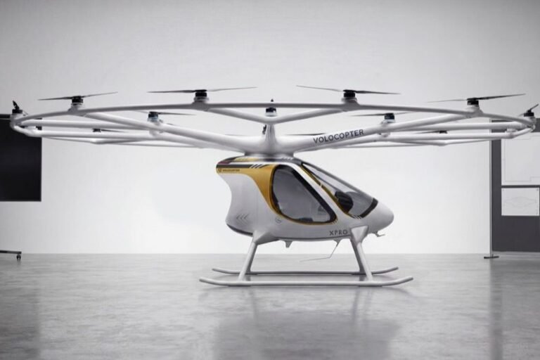 Volocopter VoloXPro two-seat ultralight electric multicopter in a studio setting, showing its ring of 18 rotors on radial arms above a white and gold enclosed cabin with skid undercarriage
