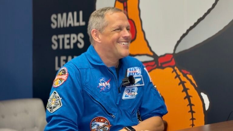 A man in a blue jumpsuit sits smiling in front of a wall with a giant snoopy astronaut painted on.