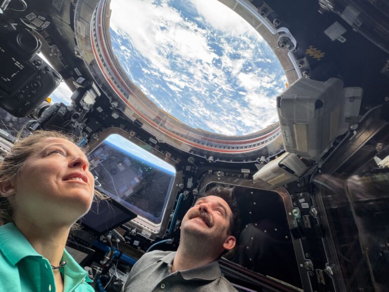 Look Up! – NASA Two astronauts look up through a round window in the International Space Station's cupola. Earth's blue water and white clouds are visible. In a trapezoid-shaped window between the two astronauts, we can see the blue haze of Earth's atmosphere.
