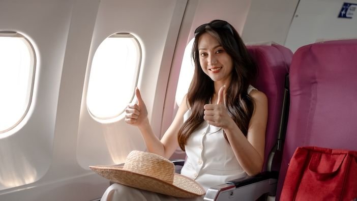 Survey highlights the US airlines with the best customer experience Smiling woman passenger giving thumbs up while seated by airplane window in economy cabin, travel hat on lap and red bag nearby, bright daylight, vacation and air travel concept.