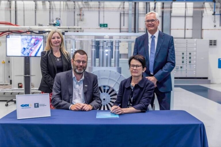 Two people sign documents at a blue-draped table bearing GKN Aerospace and University of Nottingham logos, with two colleagues standing behind and a large electrical machine test rig in a transparent enclosure in the background