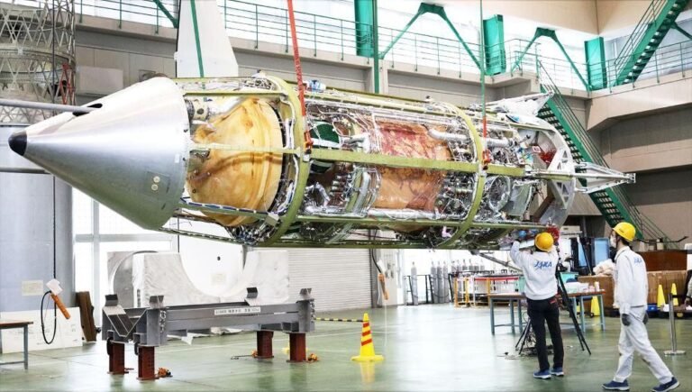 The cylindrical RV-X test vehicle lying horizontally in an assembly hall with its outer panels removed to expose internal propellant tanks, plumbing and the engine bay, a conical nose at left, as two technicians in hard hats work alongside