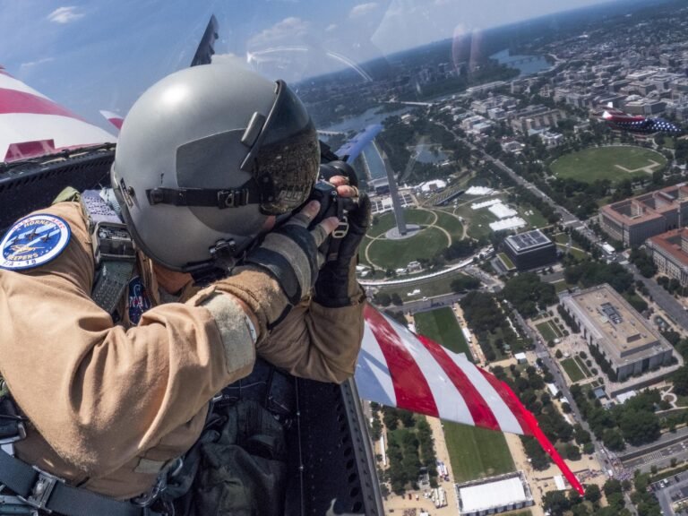 NASA Photographer Captures Images from F-18 Over Washington A man in a tan flight suit, flight gear, and helmet takes a photo with a camera over his left shoulder inside of a jet aircraft. The red and white wings of the aircraft are seen to the sides of the man. A second jet aircraft with red, white and blue paint is seen to the right of the frame. There are buildings and grassy areas below the two aircraft.