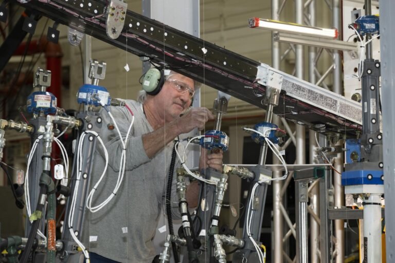 NASA Pushes New Wing Design to Find Structural Limits A man wearing ear protection works closely with multiple hydraulic and instrumentation units connected to a large beam mounted on a test structure. Numerous cables, hoses, and measurement devices extend from the setup.