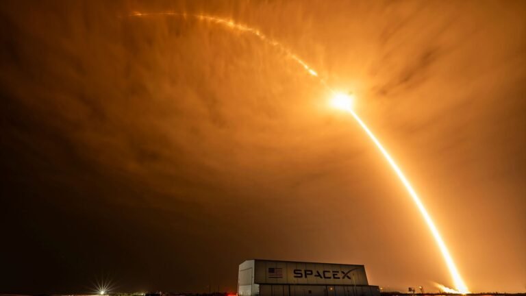 SpaceX launches flight-proven rocket for 600th time, sending Starlink satellites to orbit time lapse photo of an early morning rocket launch producing a bright streak arcing over a hangar and stretching beyond the clouds