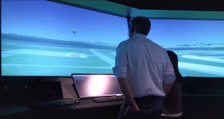 Two people standing at a workstation watching a wide curved screen that displays a simulated airport, with an autonomous aircraft in flight and another on the runway