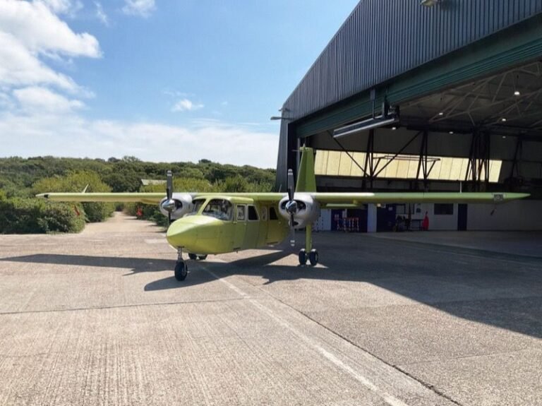 A lime-green twin-propeller Britten-Norman Islander parked on tarmac outside a hangar under clear skies