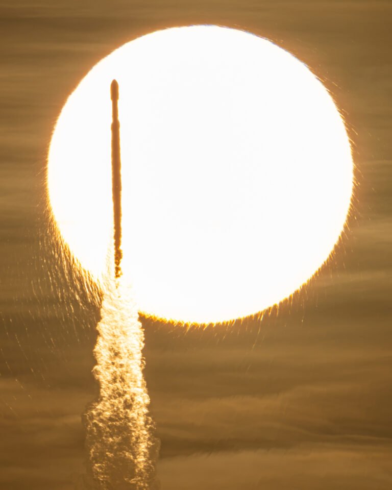 In a sepia-toned photo, the Sun appears as a bright, enormous orb that appears to have frayed edges. Against this backdrop, a tall and slender rocket is seen in silhouette atop a cloudy plume.