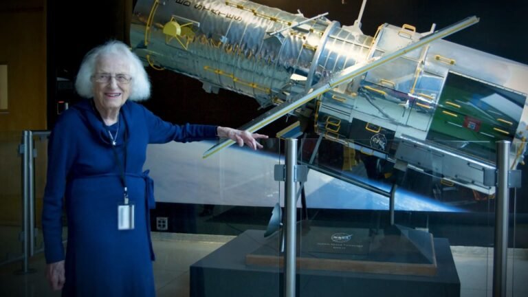 an elderly white-haired woman in a blue dress stands next to a model of a silver space telescope