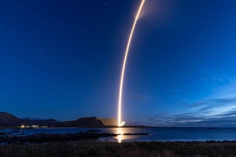 Europe’s Isar Aerospace delivers satellites to orbit on second flight Alt text: A long-exposure night photograph of the Spectrum rocket rising from the Andøya coast, its bright arcing exhaust trail reflected in the water below against a deep blue twilight sky