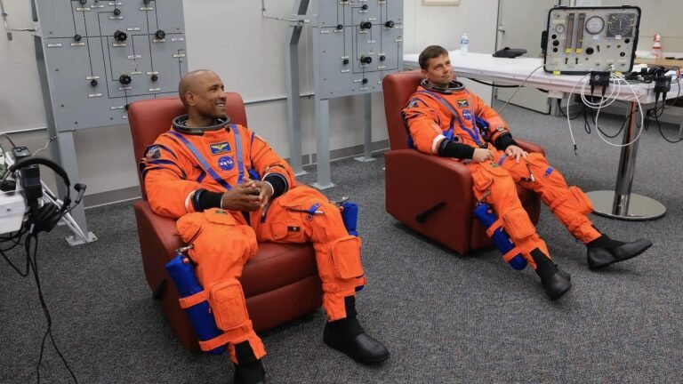 What comes after the moon? Artemis II commander and pilot shift to ’emeritus’ status at NASA two male astronauts in orange flight suits sit in reddish recliners inside a white-walled room