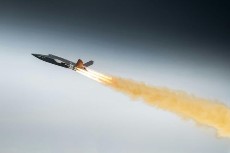 Dark grey uncrewed combat aircraft climbing steeply with a bright rocket booster flame and a long orange smoke trail against a grey sky