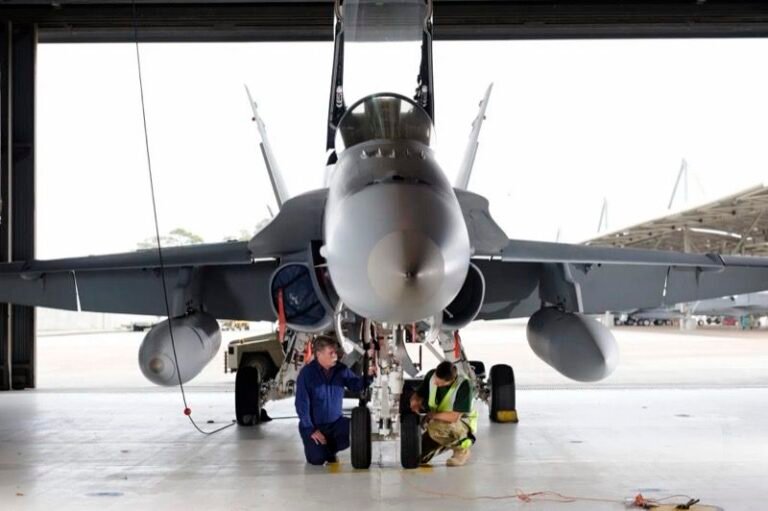 Australian project aims to certify laser repairs for fighter jets Two technicians kneel to work on the nose landing gear of an F/A-18 fighter jet in a hangar, with the aircraft’s canopy raised