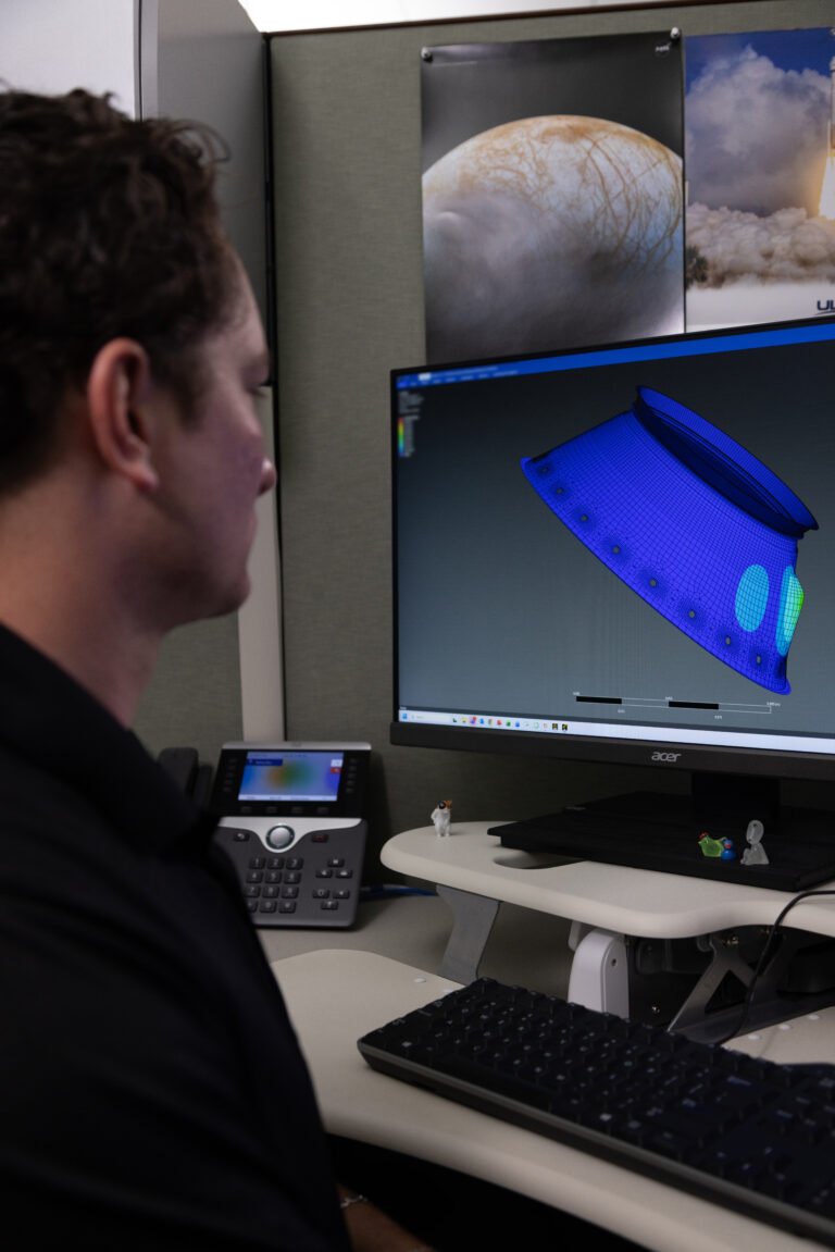 NASA Improves Shock, Vibration Analysis A person sits at a workstation viewing a computer monitor that displays a blue 3D wireframe engineering model of a spacecraft component.