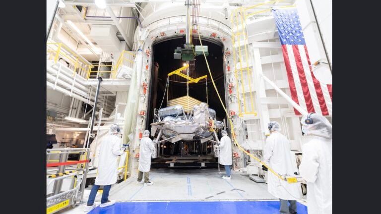 Engineers in white clean-room garb and hairnets guide a spacecraft suspended from a crane into a large thermal vacuum chamber inside a high-bay facility with an American flag hanging on the wall.
