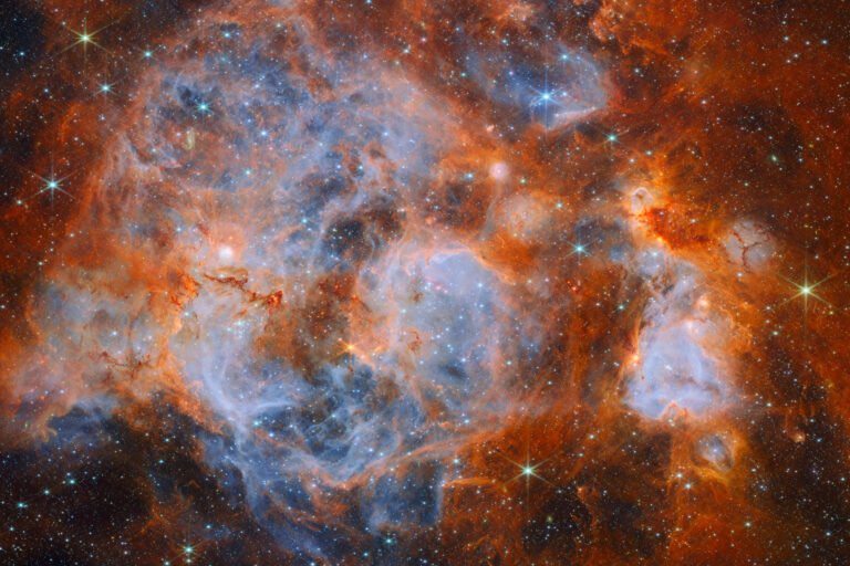 A nebula made of clouds of gas and dust with stars forming within. In the center and out to the left it is filled with thin, wispy blue gas that forms delicate layers. Around this are thicker orange clouds with dark ridges. Thin red filaments of gas cross the center of the nebula. Glowing bubbles in the gas contain dense, very bright clusters of stars. In the edges, distant stars are visible as bright points on a dark background.